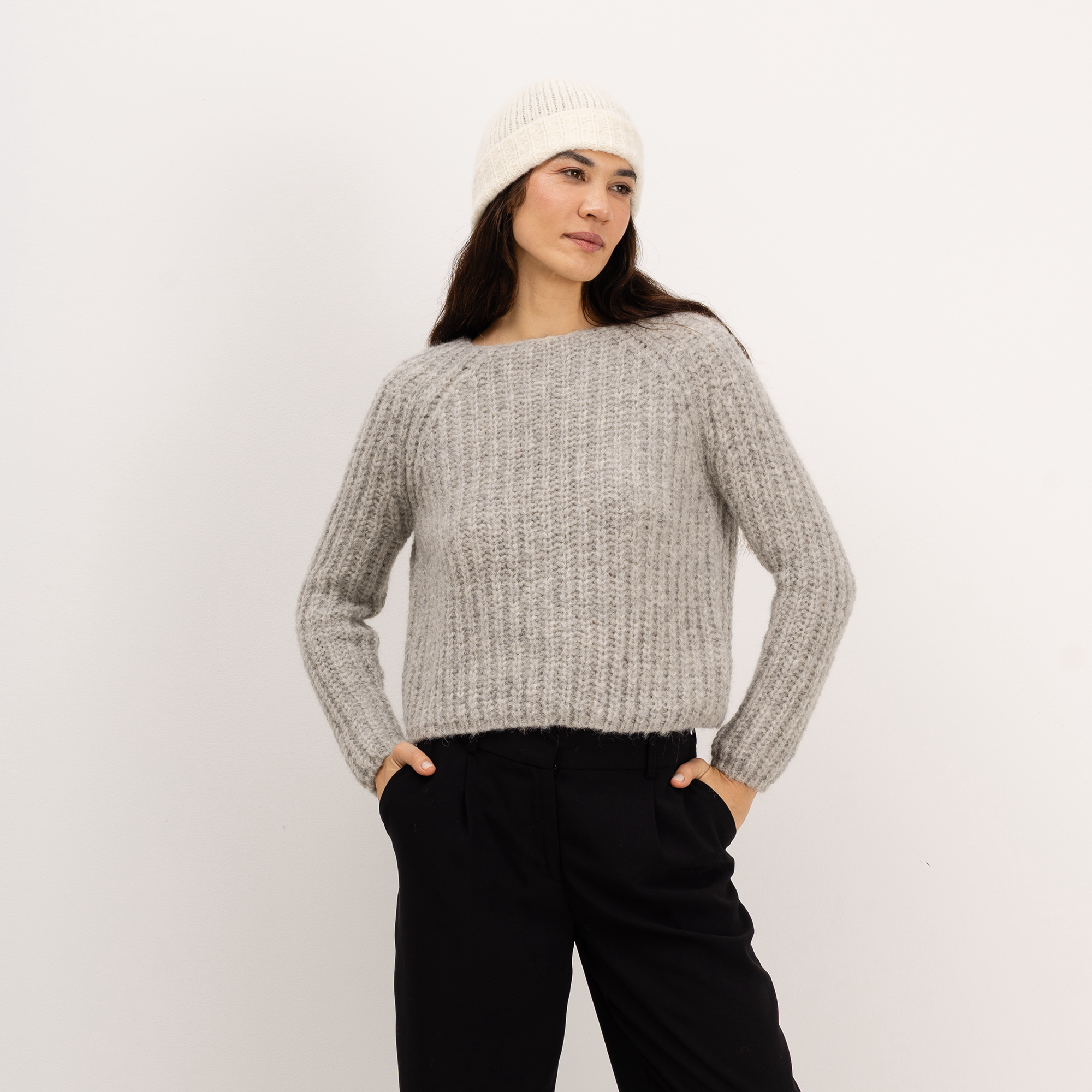 A woman wearing a light gray ribbed knit sweater, black pants, and an off-white beanie hat, standing against a plain light gray background.