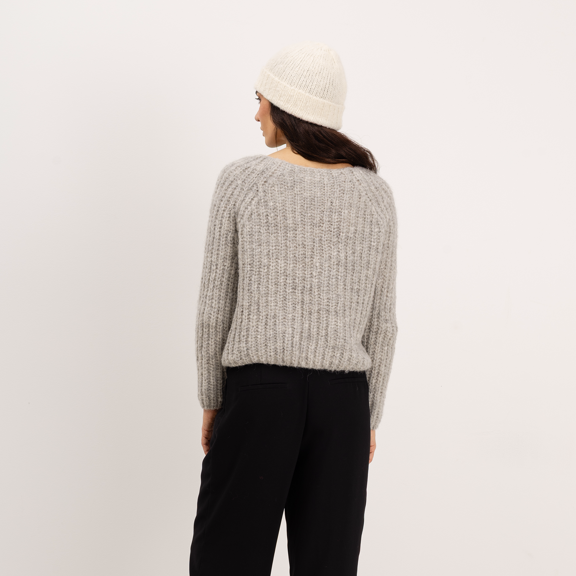 A woman shown from the back wearing a light gray ribbed knit sweater, a cream beanie, and black pants, standing against a plain light gray studio background.