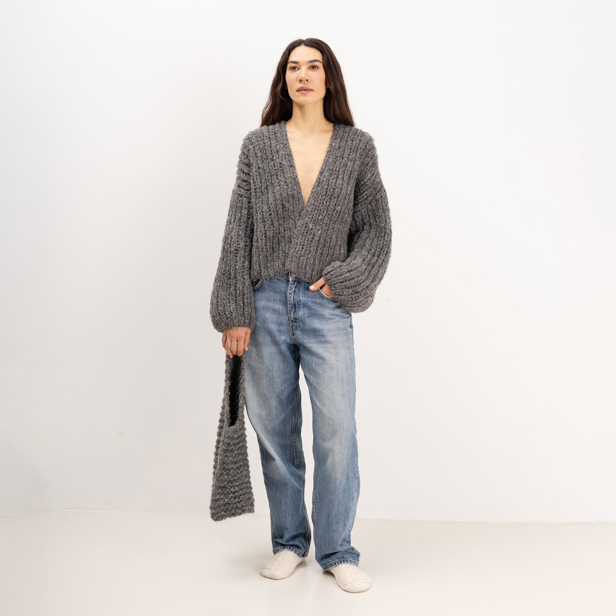 A woman stands against a plain white studio background wearing a chunky gray knit wrap cardigan with balloon sleeves, light-wash blue jeans, and white shoes, while holding a matching gray knit tote bag.