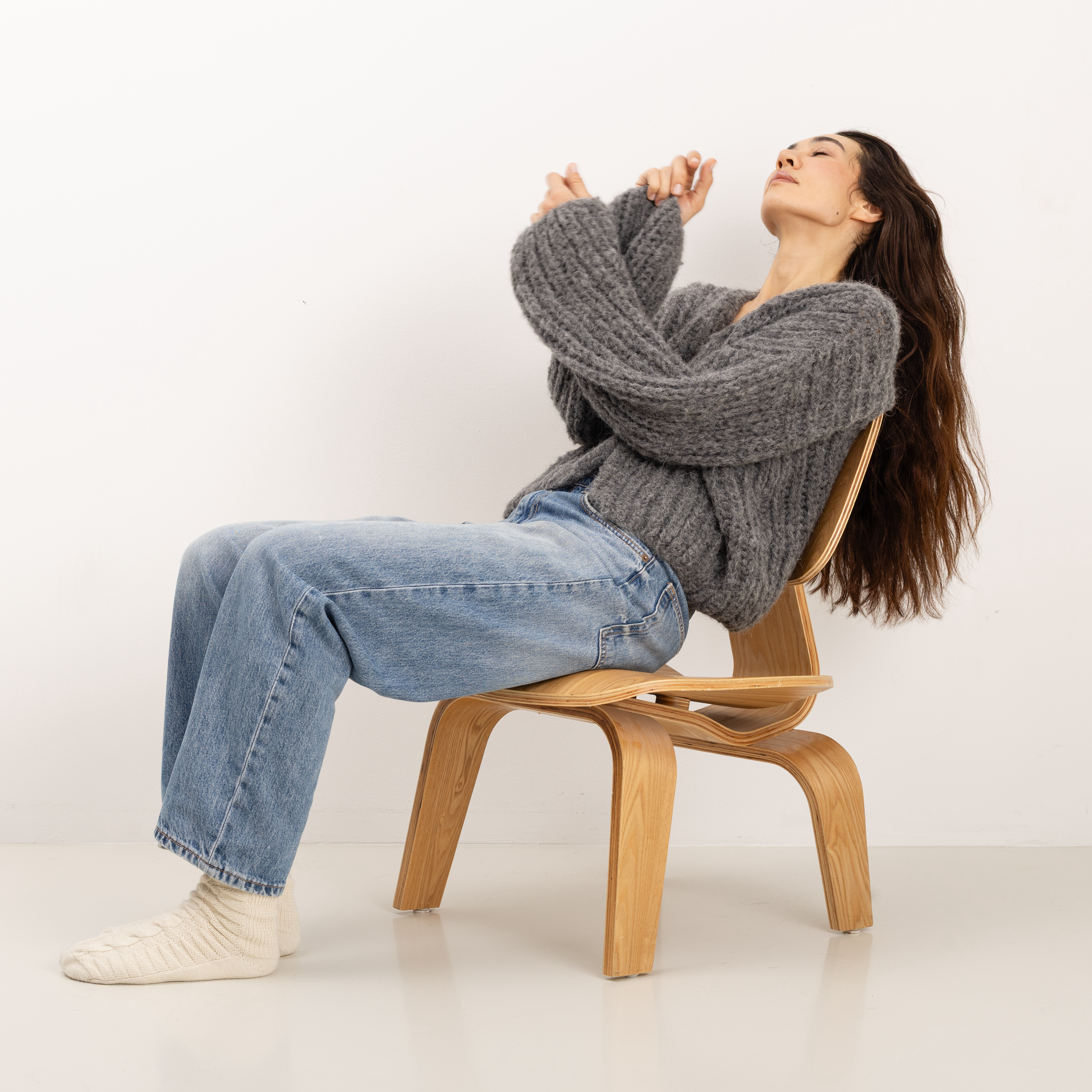 A woman with long dark hair sits on a light wood chair, leaning back with her arms crossed over her chest. She is wearing a chunky gray knit sweater, light blue jeans, and cream-colored socks against a plain white studio background.