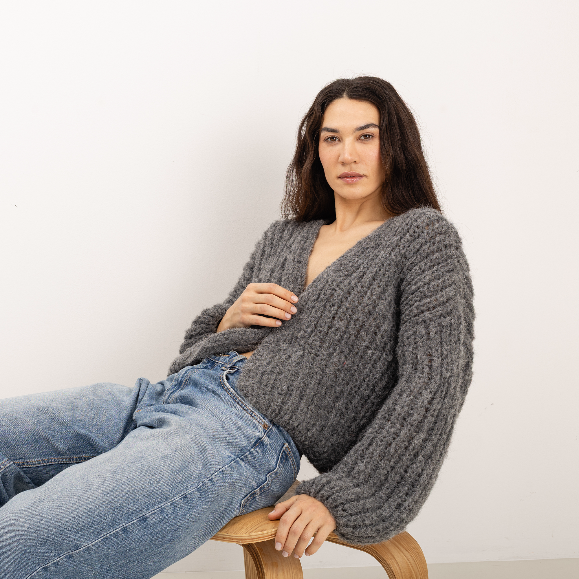 A woman wearing a chunky gray cable-knit V-neck sweater and blue jeans, sitting on a wooden stool against a plain off-white wall.