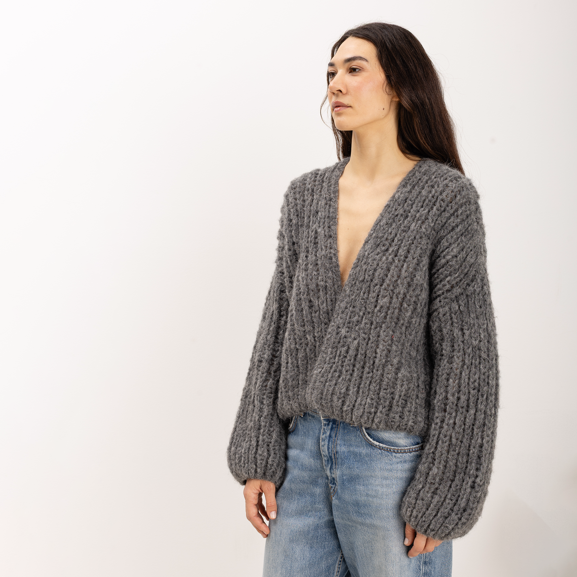 A woman wearing a chunky gray ribbed knit cardigan sweater with a deep V-neck and long balloon sleeves, paired with light blue jeans, standing against a plain white studio background.