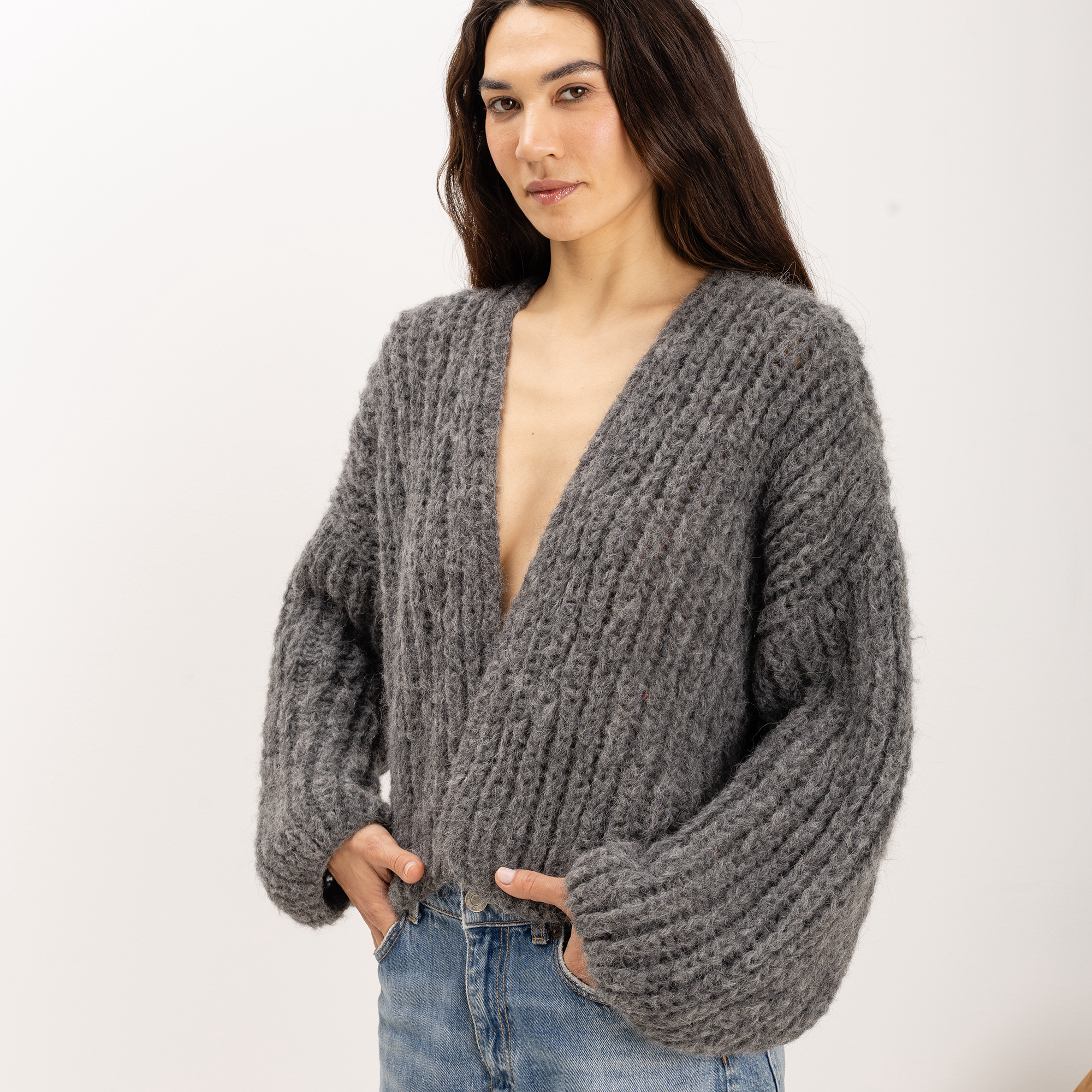 A woman wearing a chunky gray cable-knit cardigan sweater with a deep V-neck and oversized sleeves, paired with blue jeans, against a plain white studio background.