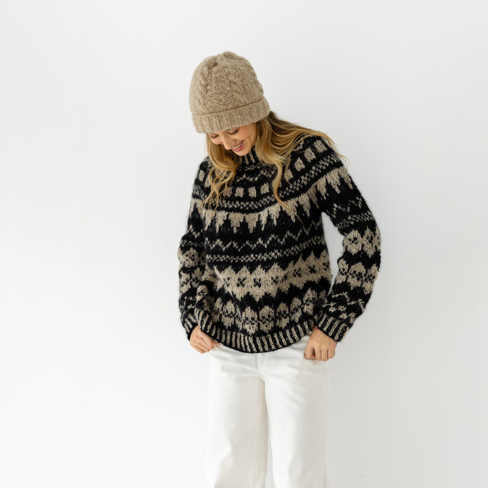 A woman wearing a black and beige patterned knit sweater, a beige beanie, and white pants, standing against a plain white background.