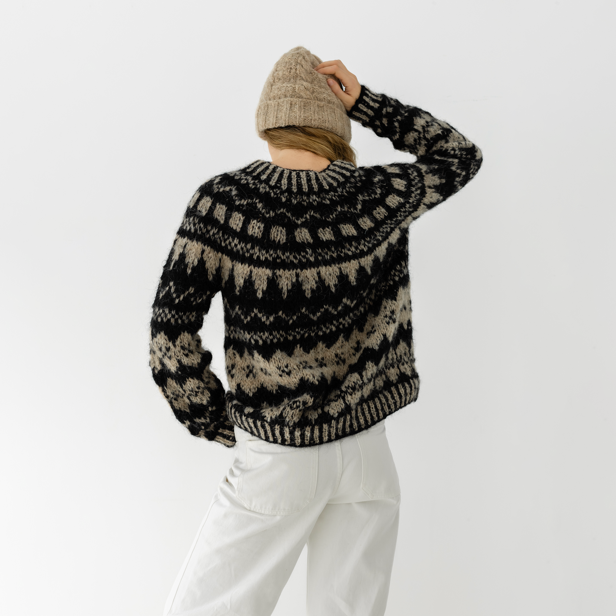 A person shown from the back wearing a black and beige patterned knit sweater, a beige beanie, and white pants against a plain light gray studio background.