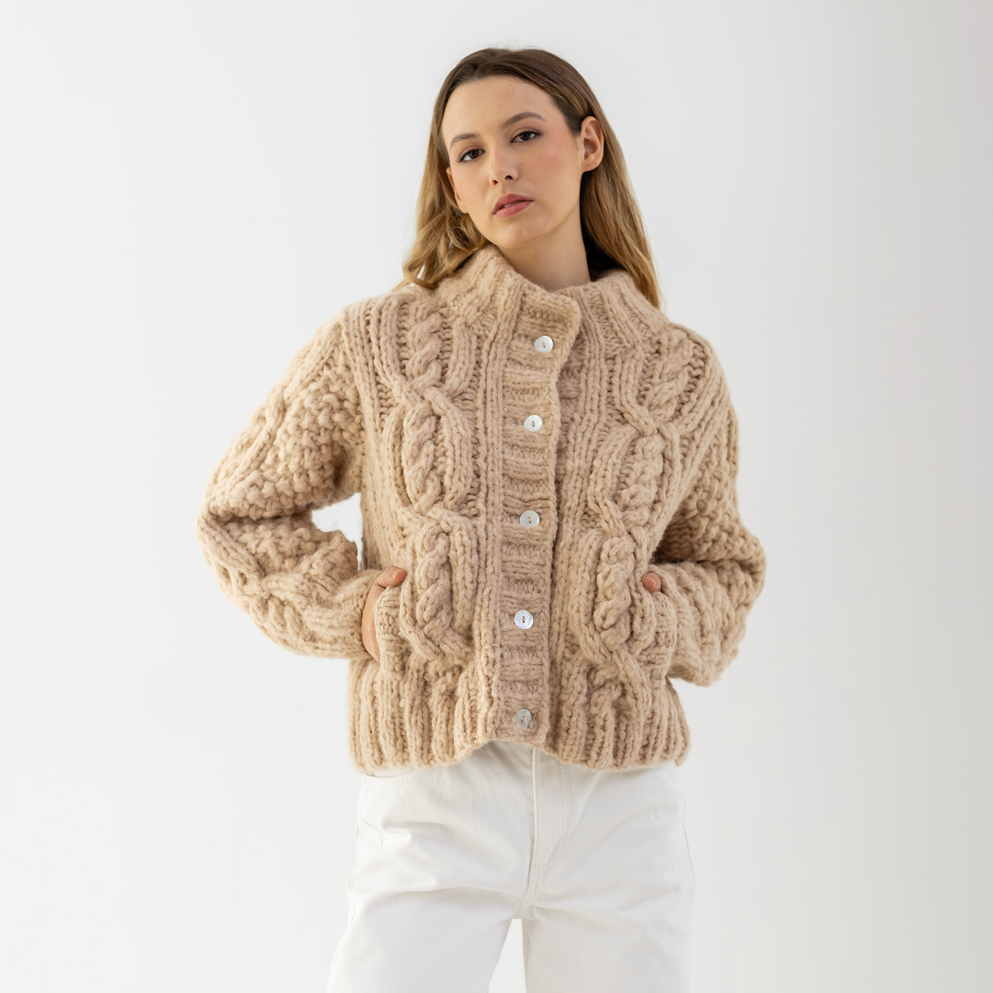 A woman wearing a chunky cable-knit cropped cardigan in a warm beige color, paired with white pants, against a plain light gray studio background.