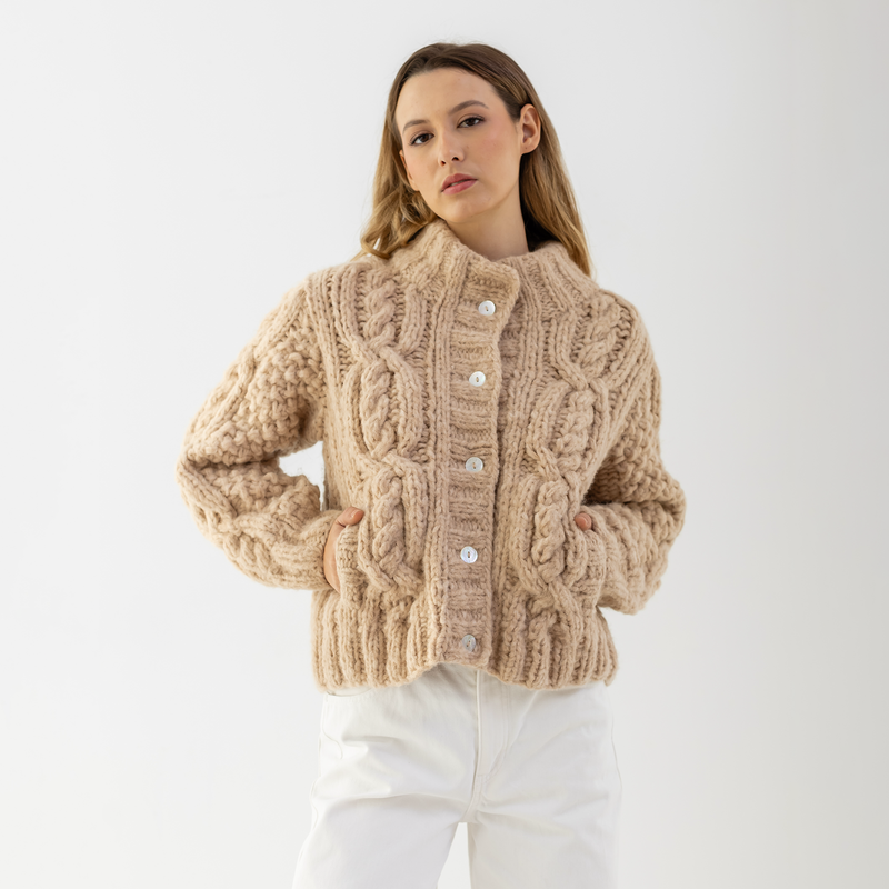 A woman wearing a chunky cable-knit cropped cardigan in a warm beige color, paired with white pants, against a plain light gray studio background.
