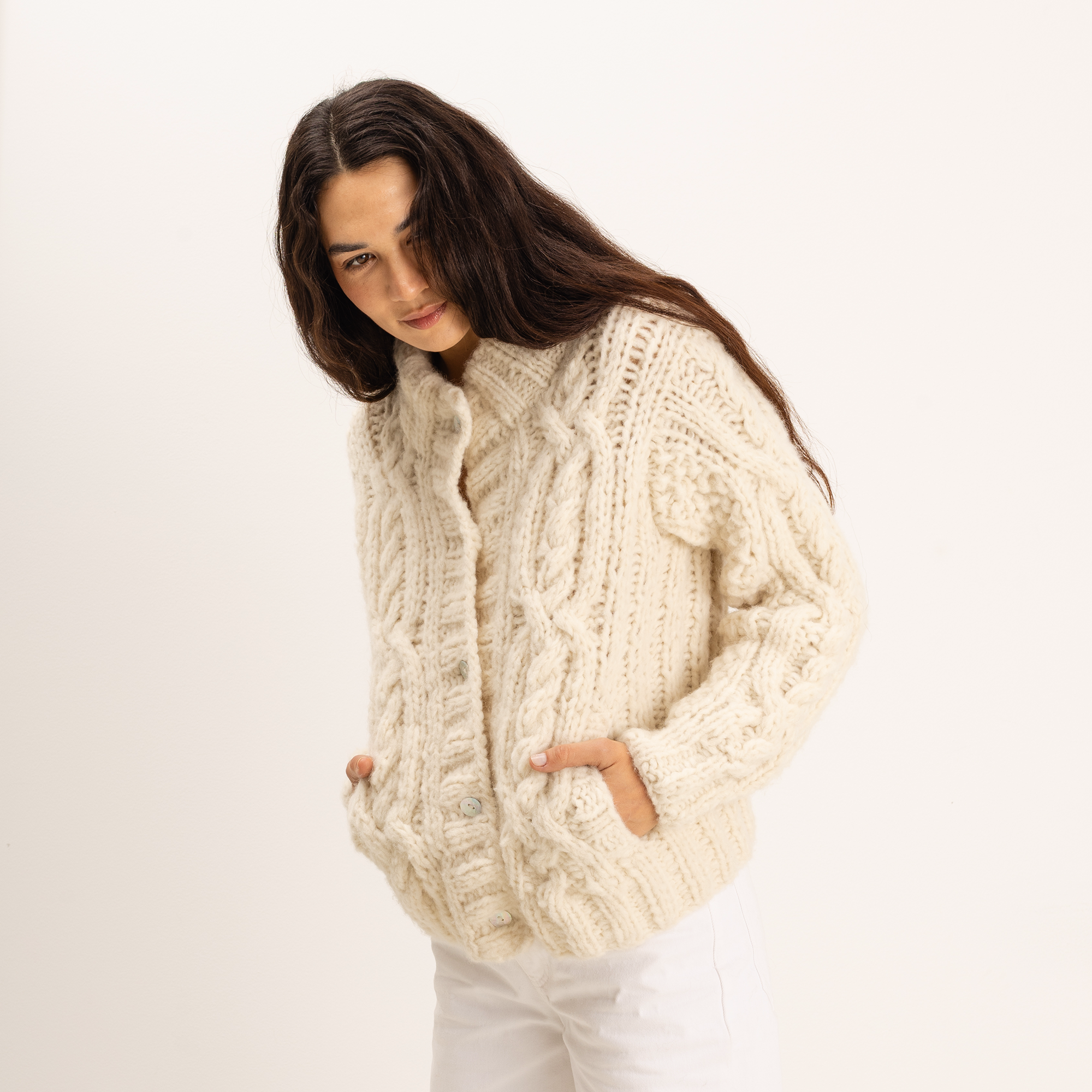 A woman wearing a chunky cream cable-knit cardigan sweater and white pants, photographed against a plain light background.