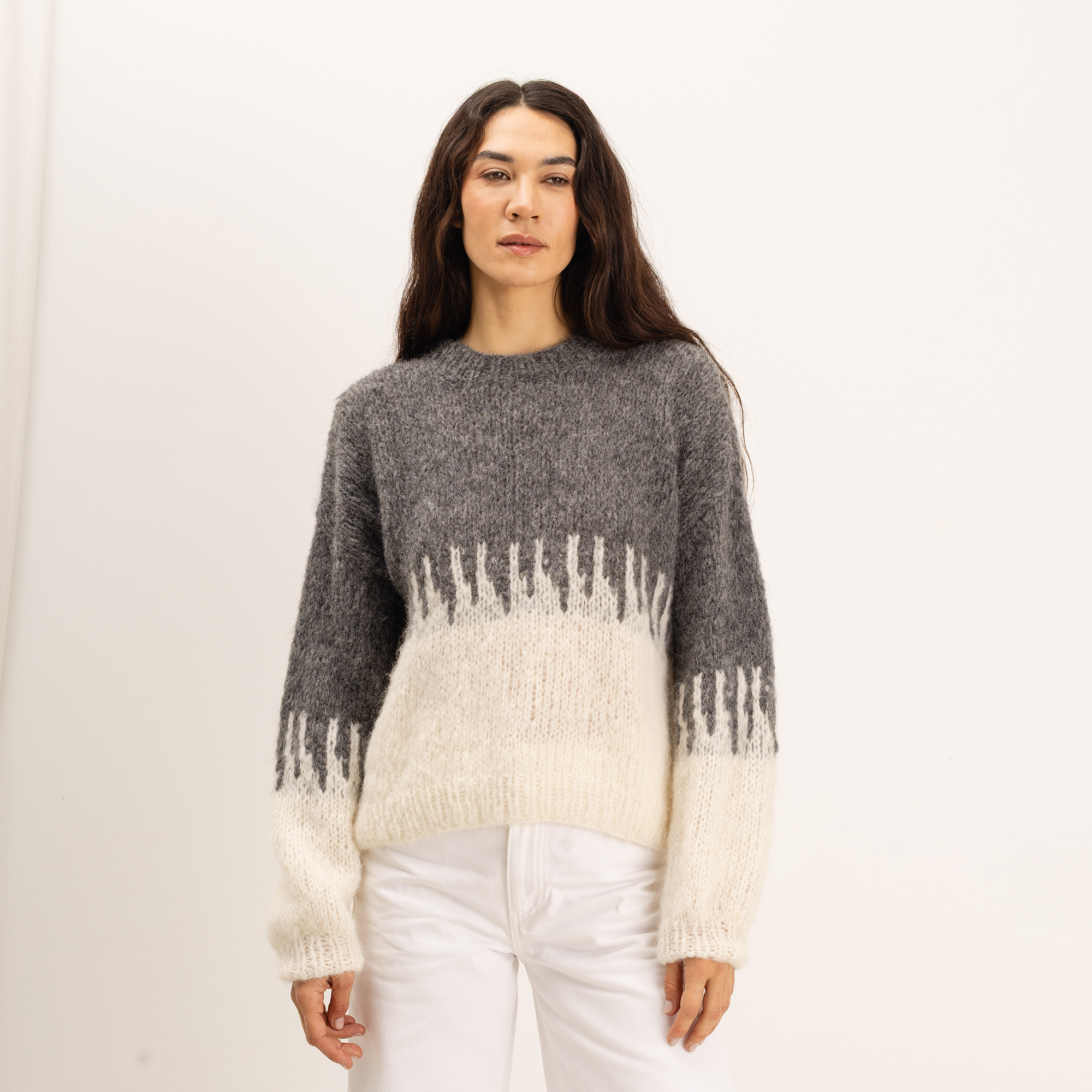 A woman wearing a fuzzy knit sweater with a gray upper section and an off-white lower section and sleeves, paired with white pants, against a plain white studio background.