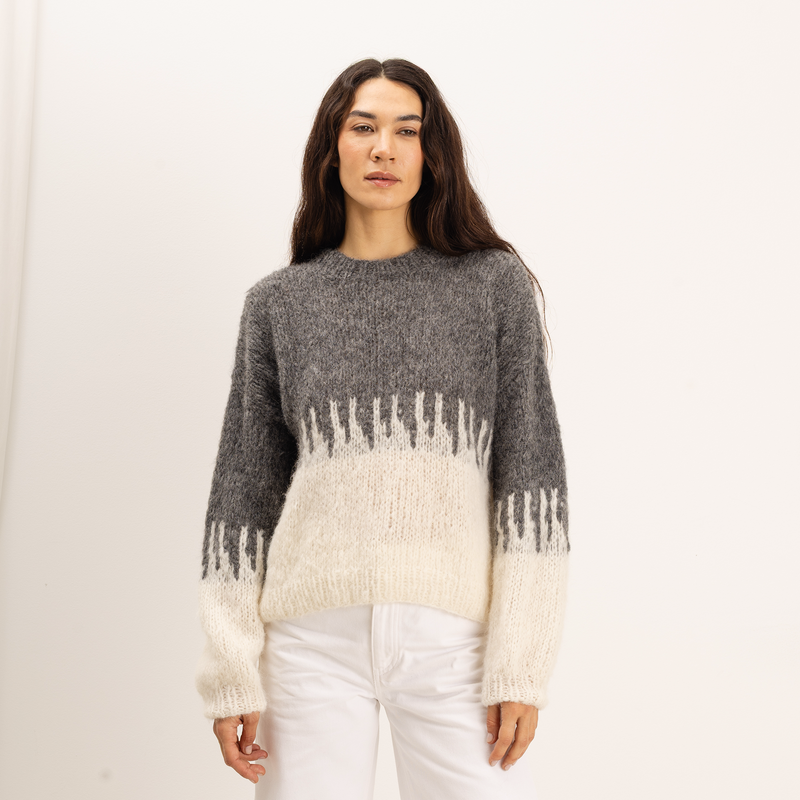 A woman wearing a fuzzy knit sweater with a gray upper section and an off-white lower section and sleeves, paired with white pants, against a plain white studio background.