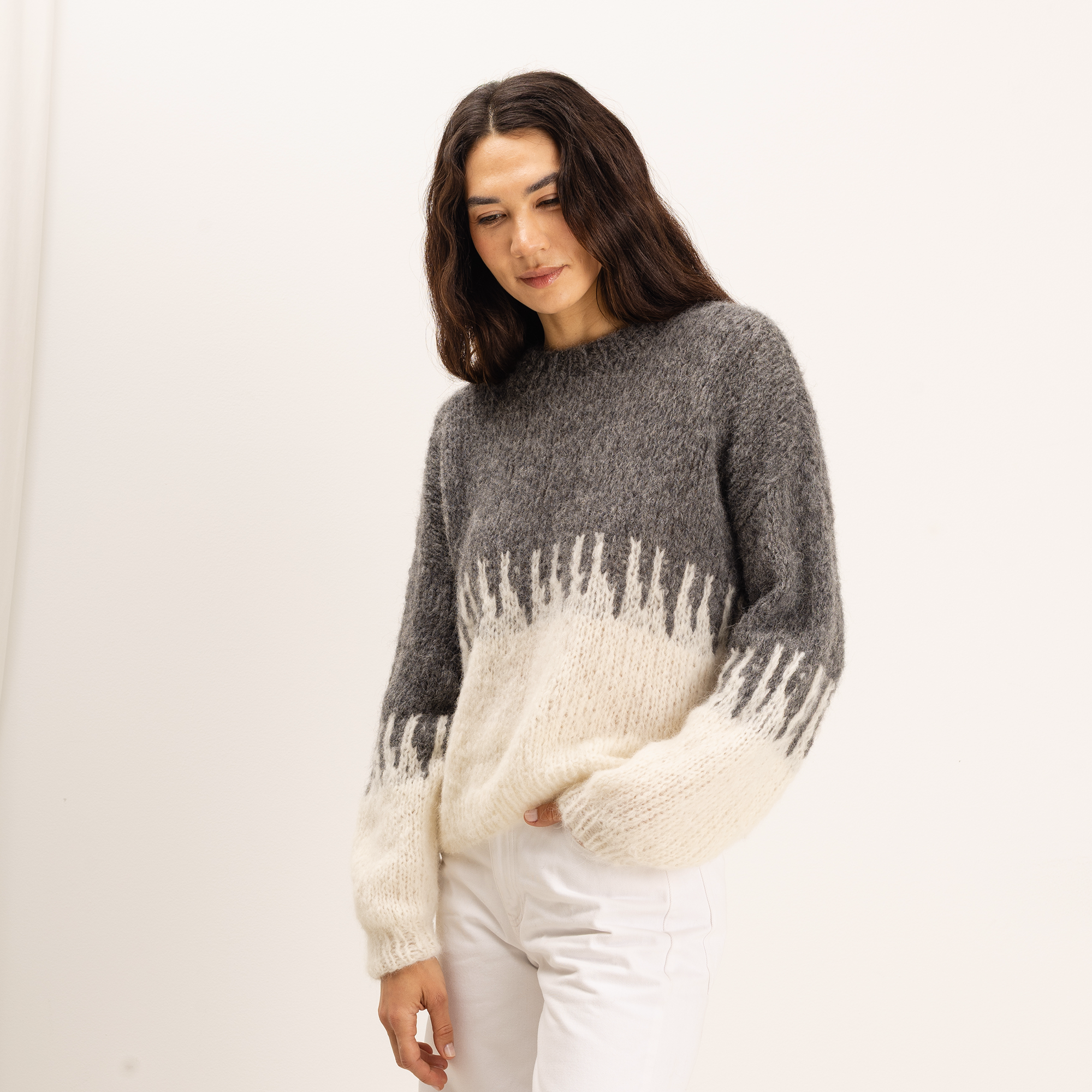 A woman wearing a fuzzy knit sweater with a gray upper body and sleeves, and an off-white lower body and sleeves with a fringe-like pattern, paired with white pants, set against a plain white background.