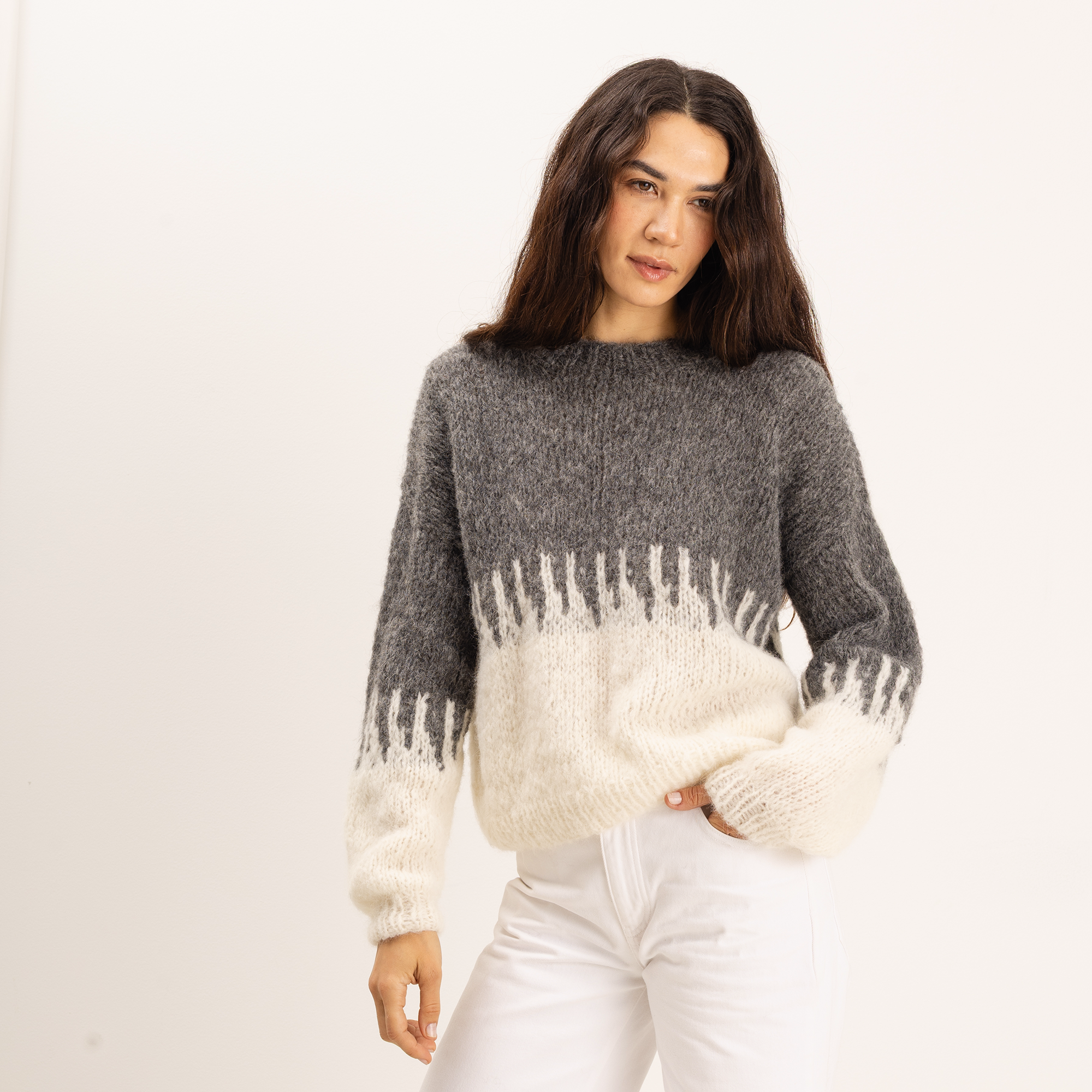 A woman modeling a fuzzy knit sweater with a color-blocked design in dark gray and cream, paired with white pants, against a plain white studio background.
