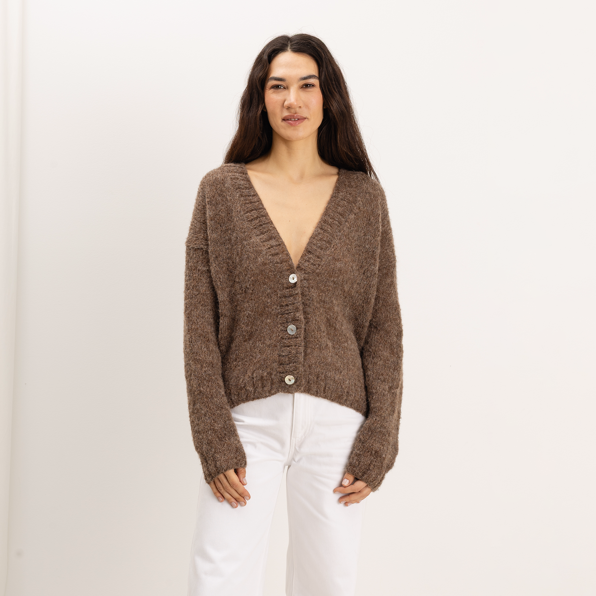 A woman modeling a brown chunky knit cardigan with a V-neck and front buttons, paired with white pants, against a plain white studio background.