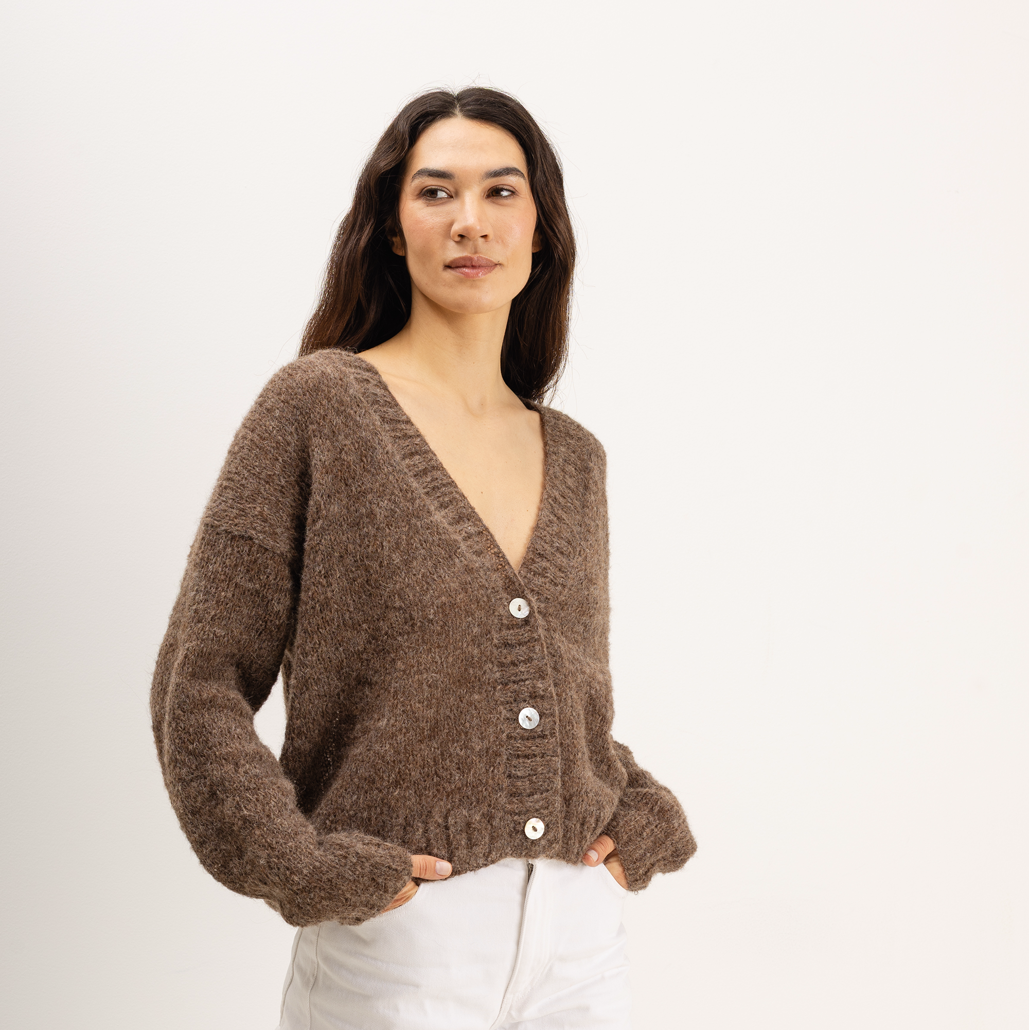 A woman wearing a brown fuzzy knit cardigan sweater with white pants, photographed against a clean off-white studio background.