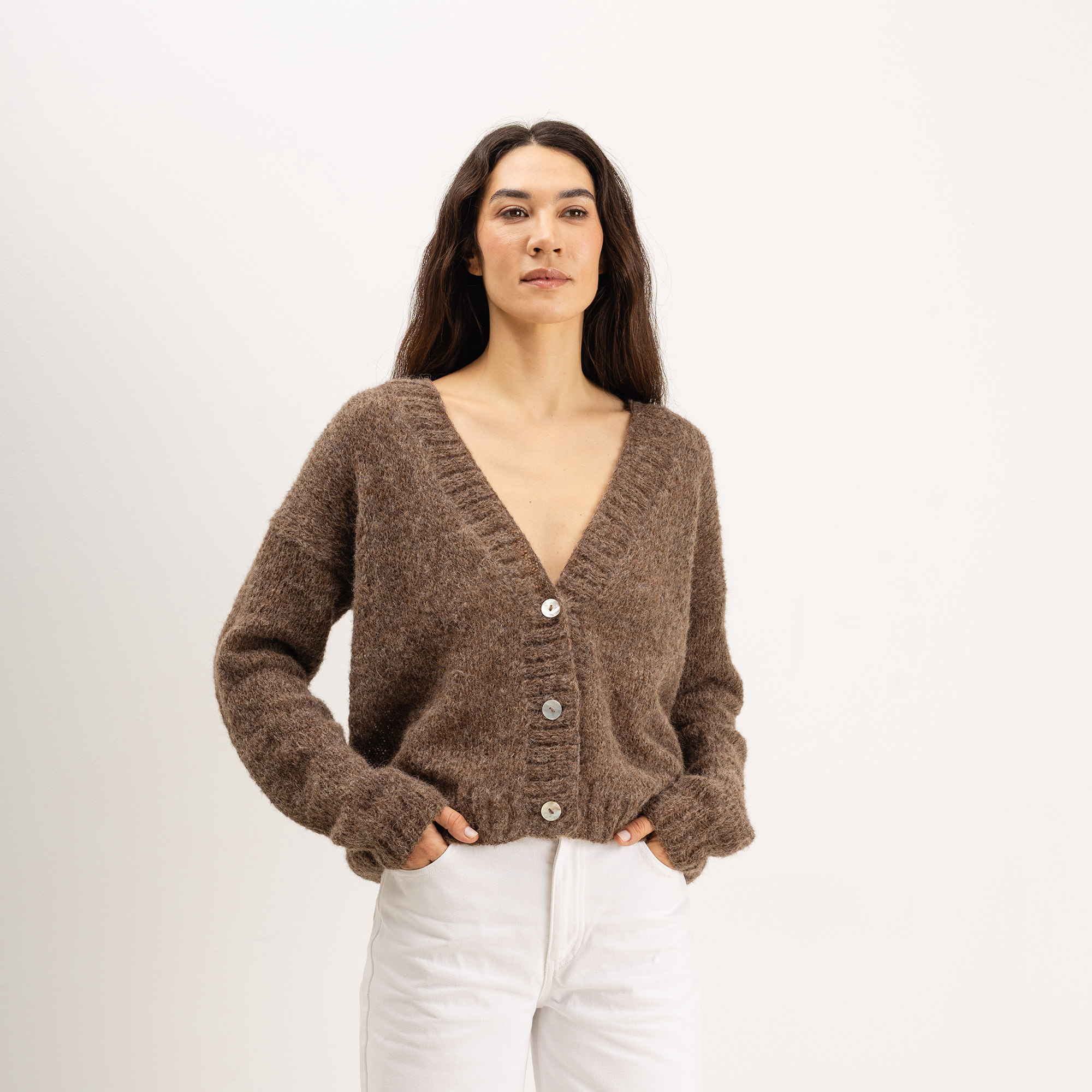 A woman wearing a brown fuzzy knit cardigan with a V-neck and silver buttons, paired with white pants, standing against a plain white studio background.
