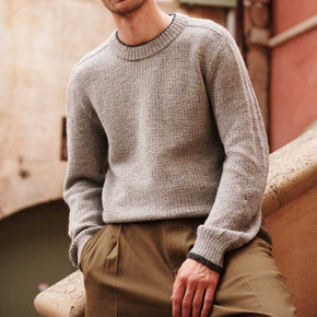 Man wearing a gray sweater and brown pants sitting on a stone ledge.