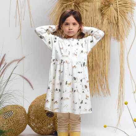 Young girl wearing a patterned dress with yellow leggings standing in front of decorative plants.