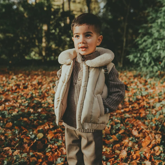 Donsje Regu Bodywarmer - child wearing light tan puffer vest with faux sherpa collar, pockets, and snap closure on wooded background