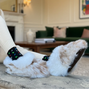Woman sitting on a stone ledge wearing fluffy white slippers with colorful bows in a cozy living room.