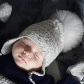 Newborn baby wearing a gray knit hat with a white pom-pom, lying on soft fabric.