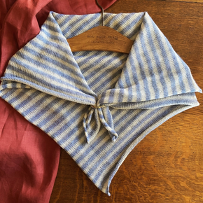 A striped alpaca wool neckerchief in blue and white tones, displayed on a wooden surface with a red garment partially visible on the left side.