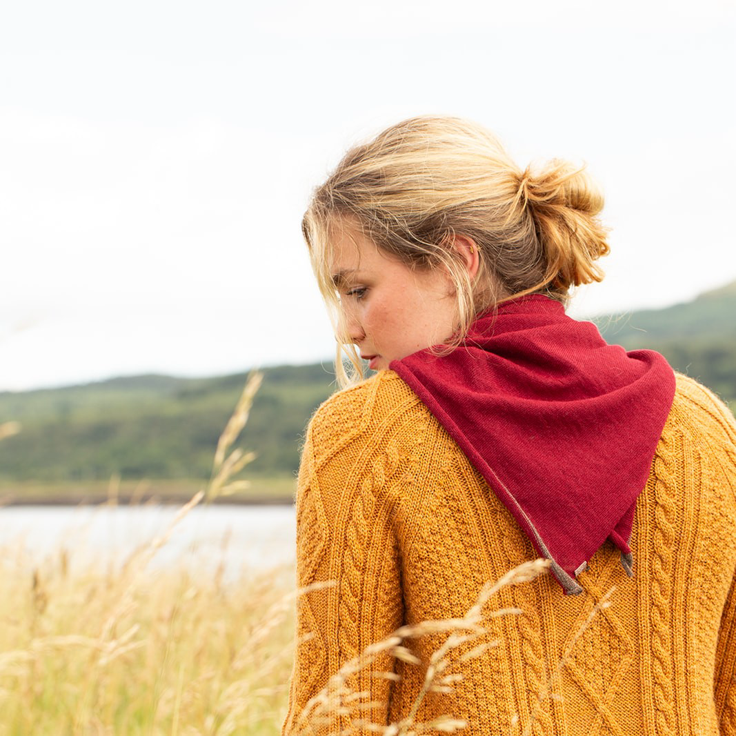 A woman in a golden yellow cable-knit sweater and a deep red neckerchief, photographed from behind in an outdoor field with a blurred lake and hills in the background.