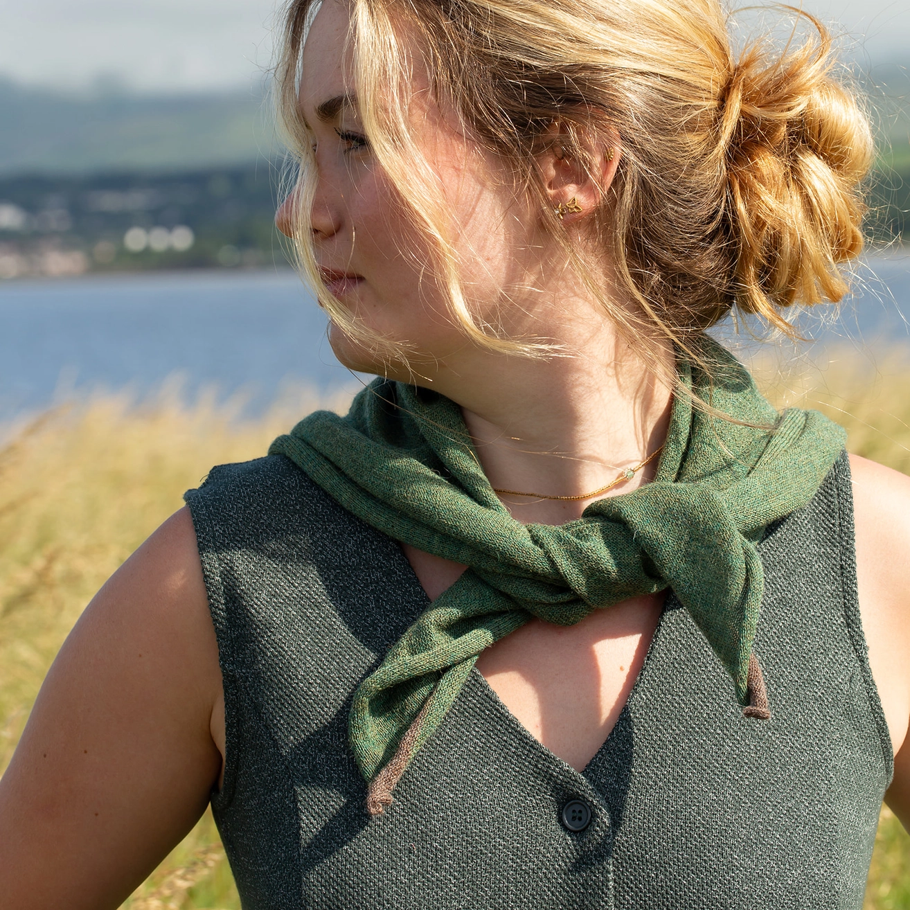 A woman outdoors wearing a dark green knit scarf tied around her neck over a dark gray sleeveless top, with a blurred lake and grassy background.