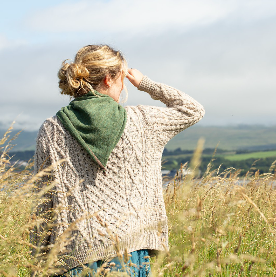 A woman seen from behind in a grassy field, wearing a cream cable-knit sweater over a green neckerchief, with a hazy landscape in the background.