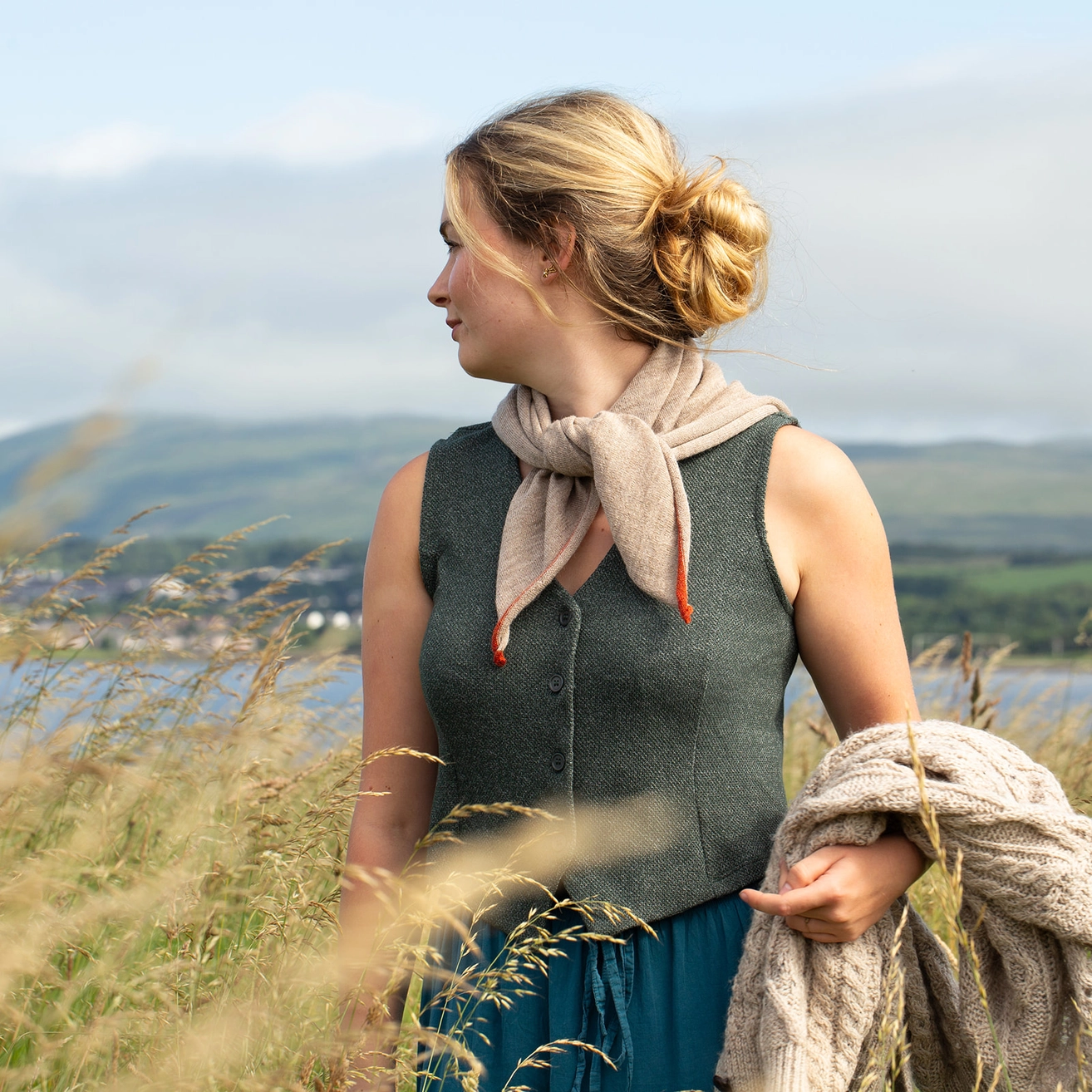 A woman standing outdoors in a grassy field with water and hills in the background, wearing a sleeveless dark gray knit vest, a beige scarf, and holding a chunky knit cream sweater.