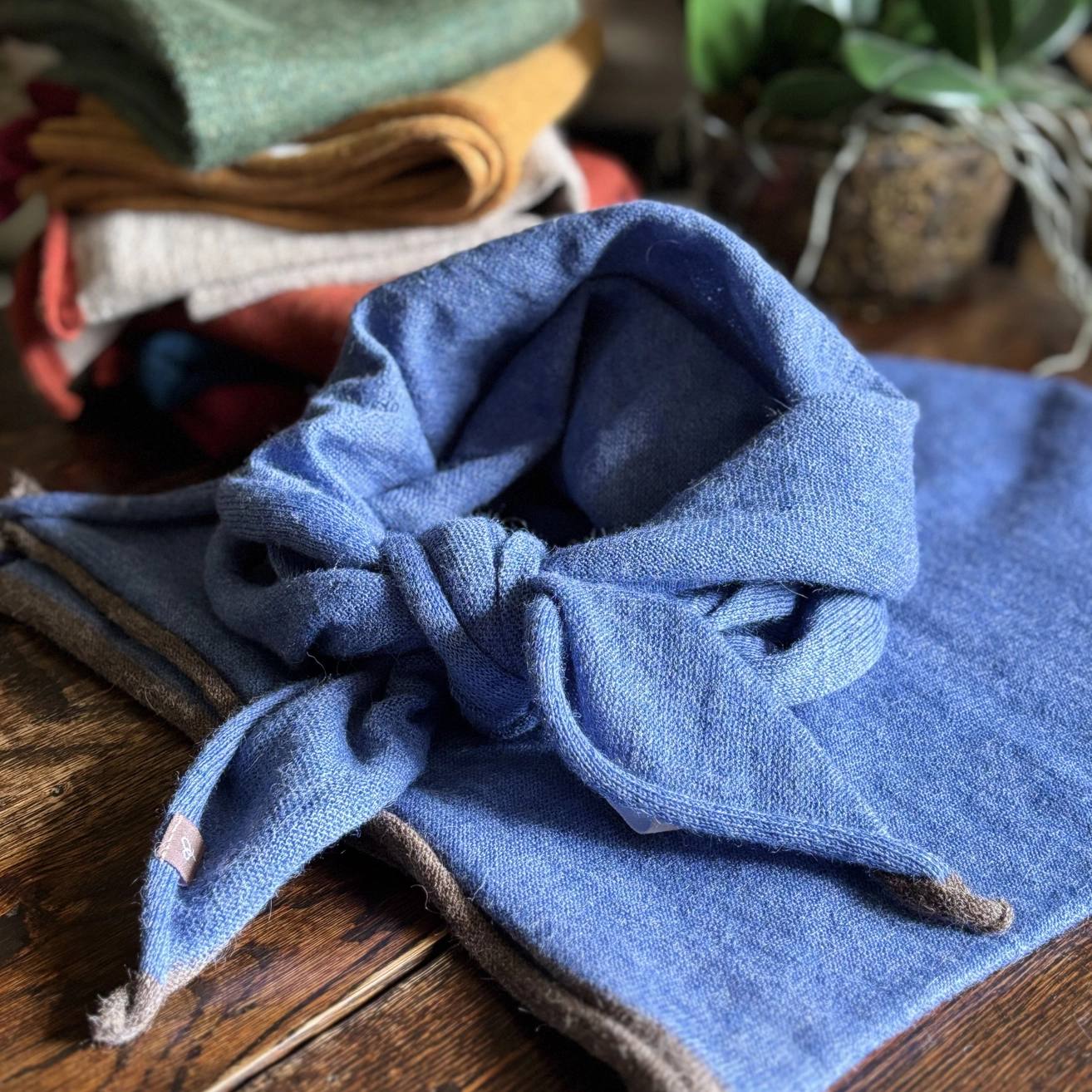 A blue knitted neckerchief tied in a knot, displayed on a dark wooden surface with other folded textiles blurred in the background.