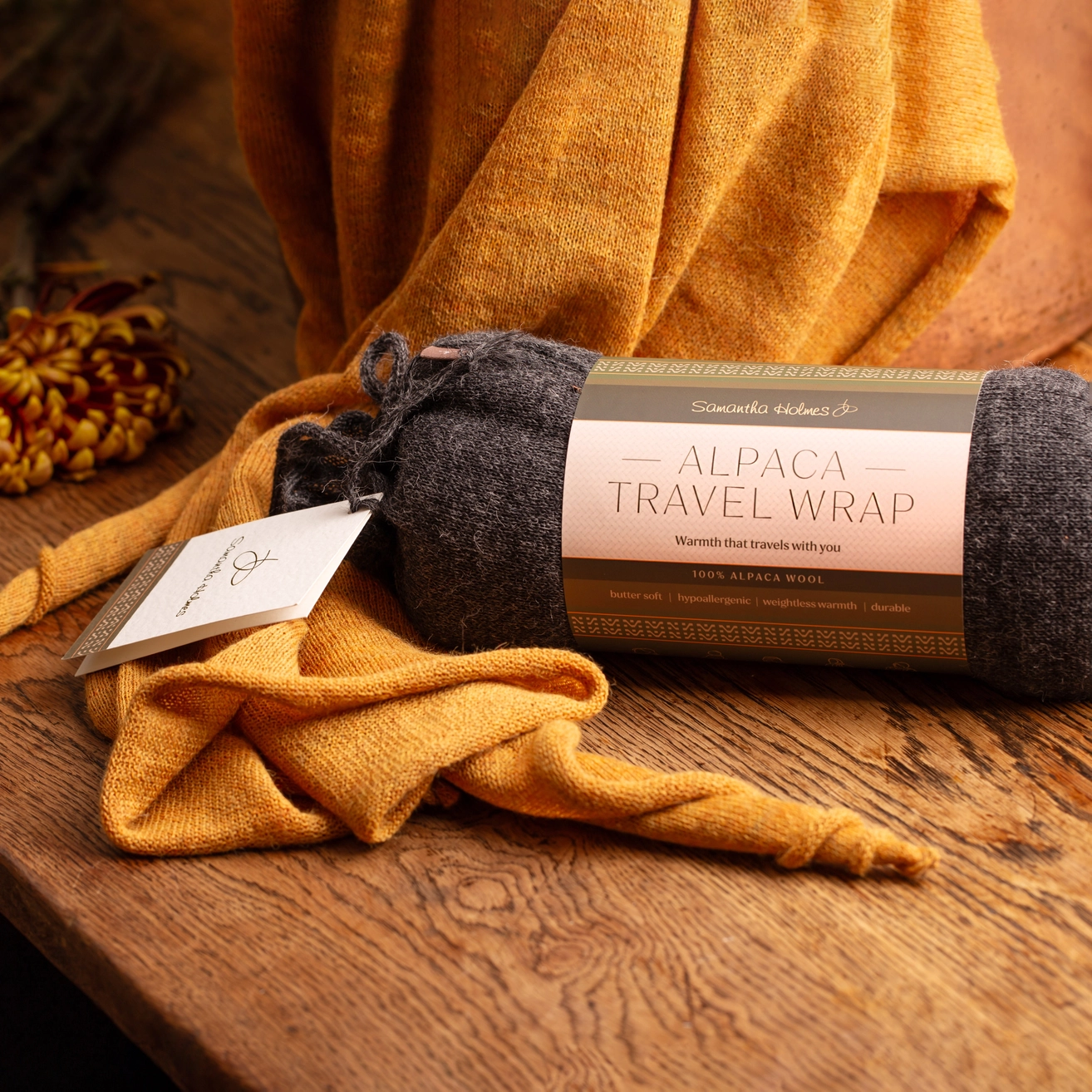A rolled Alpaca Travel Wrap in charcoal gray, wrapped in a paper band label, with a folded mustard yellow version draped around it on a rustic wooden table.