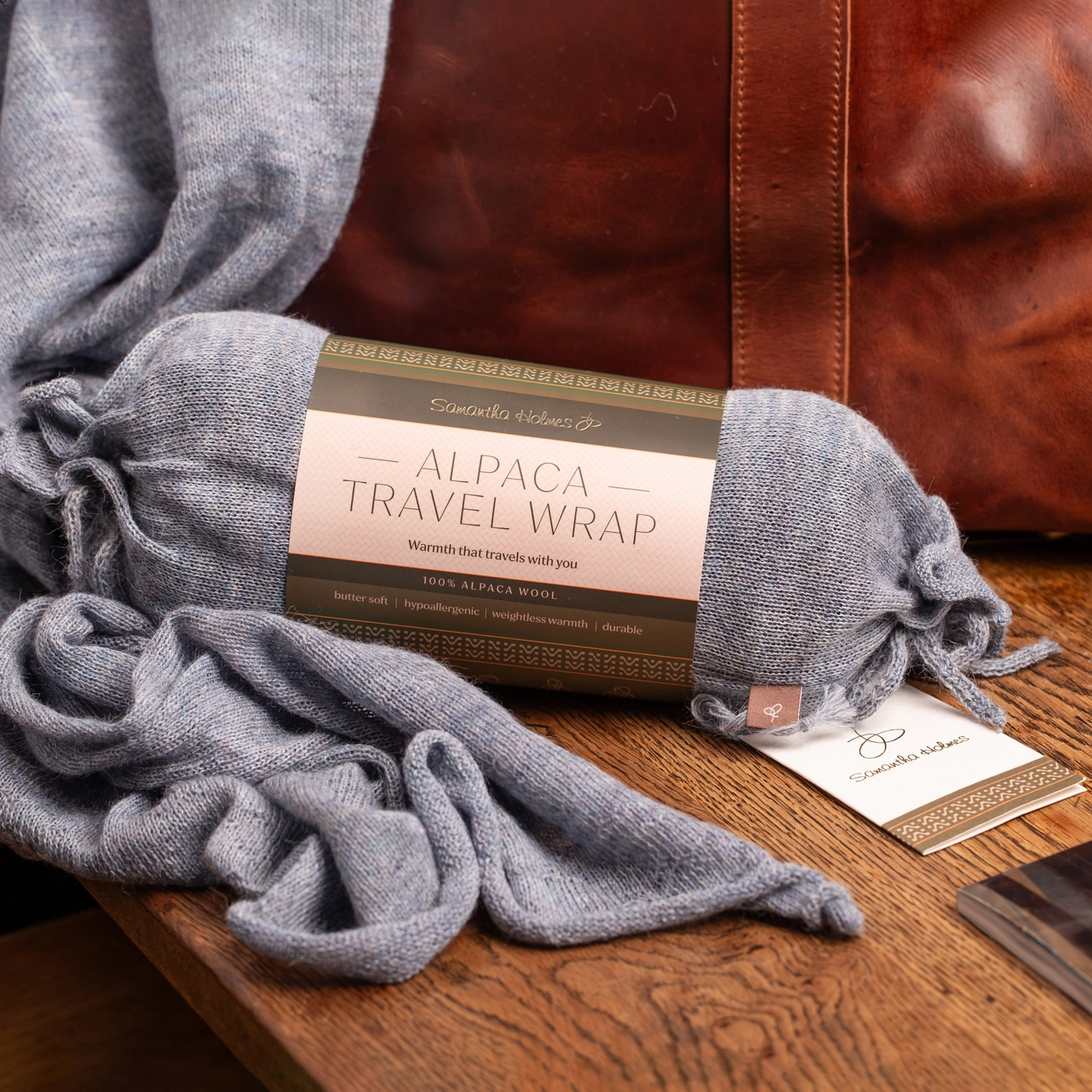 A light blue alpaca travel wrap wrapped in its paper band packaging, draped on a wooden table with a brown leather bag in the background.