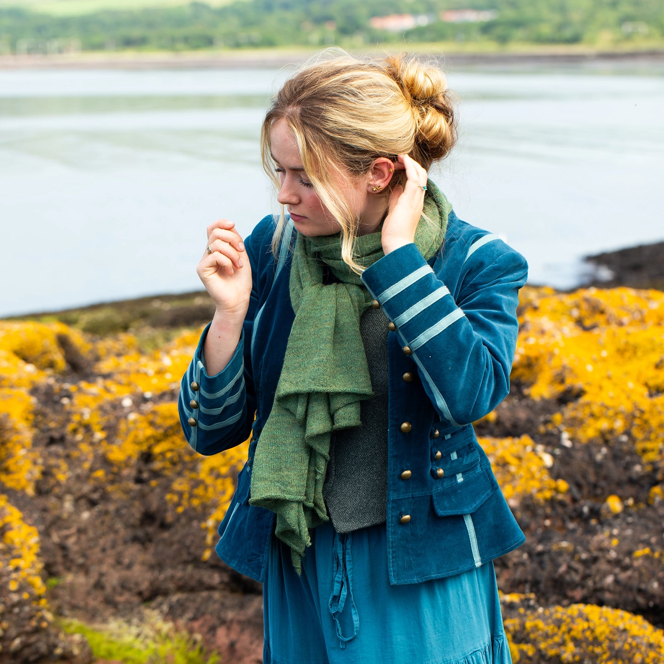 A woman standing outdoors by the water wearing a teal blue velvet jacket with white stripes on the sleeves, a green scarf, and a blue dress. Yellow coastal flowers are in the foreground.