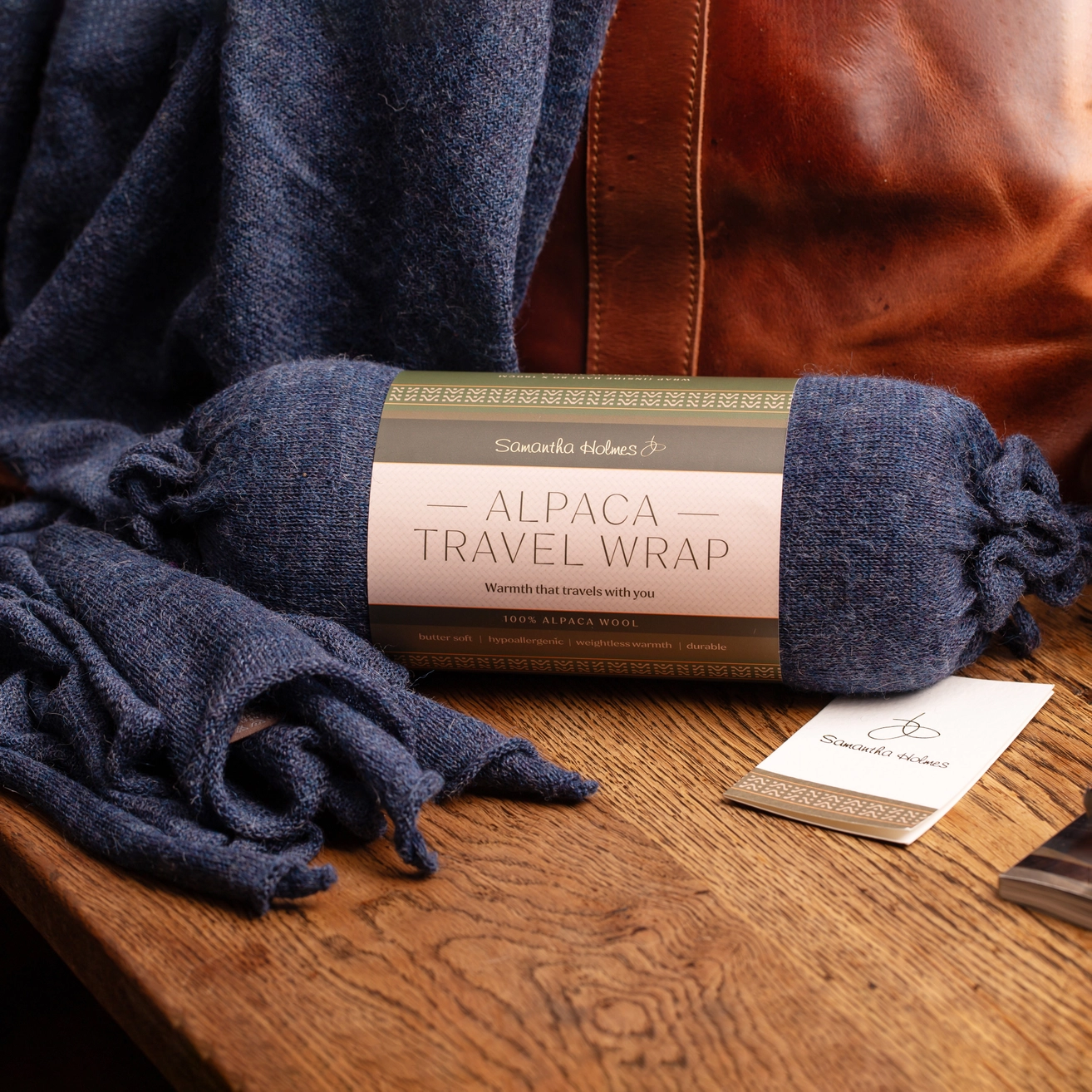 A rolled blue alpaca travel wrap with a paper label sits on a wooden table next to a brown leather chair. More of the blue wrap is draped out in front, and a small branded card is placed nearby.