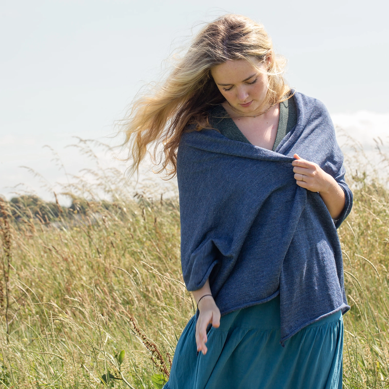 A woman standing in a grassy field outdoors, wearing a blue shawl draped over her shoulders and a teal skirt.