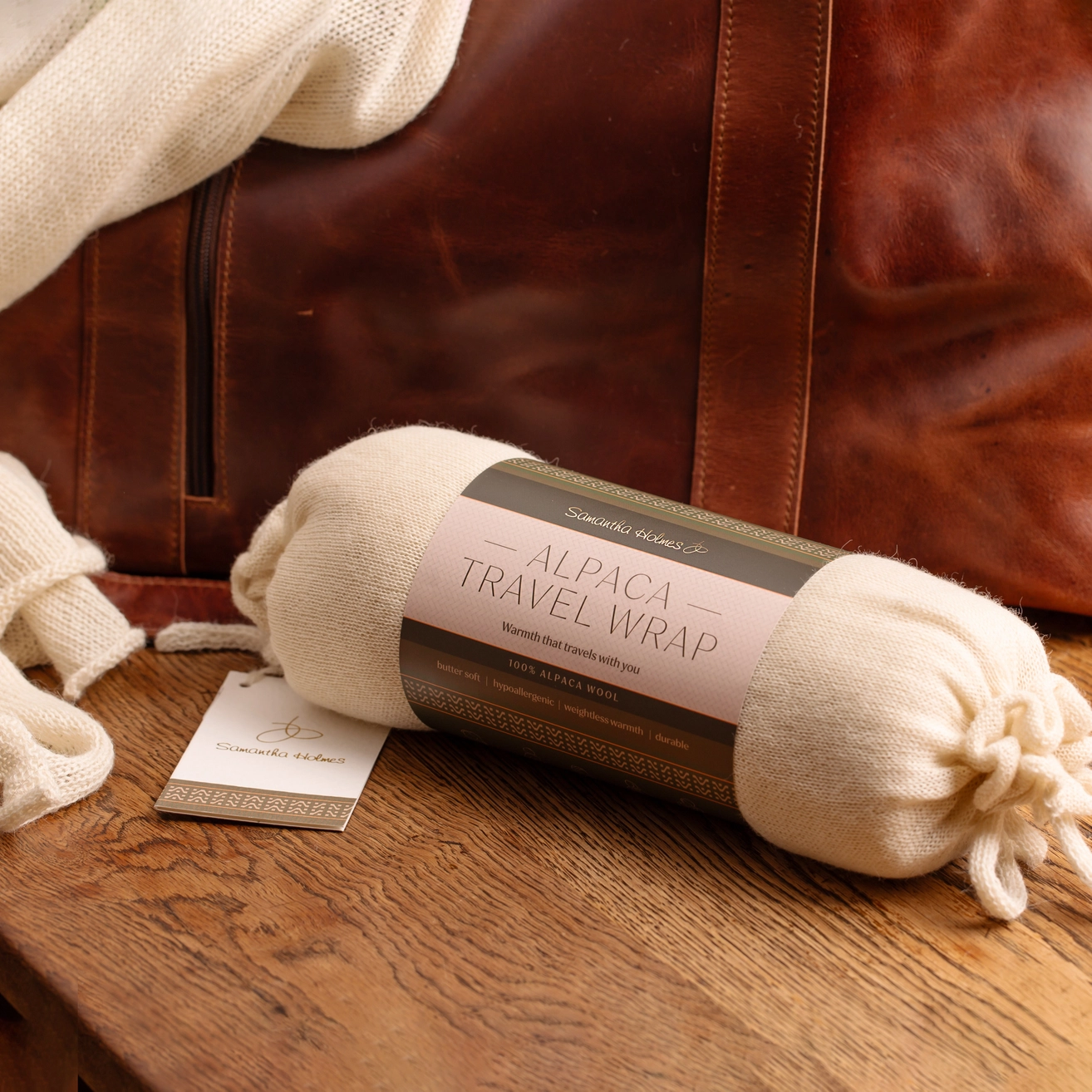 A cream-colored alpaca travel wrap rolled up with its paper label, styled on a rustic wooden table next to a brown leather suitcase.