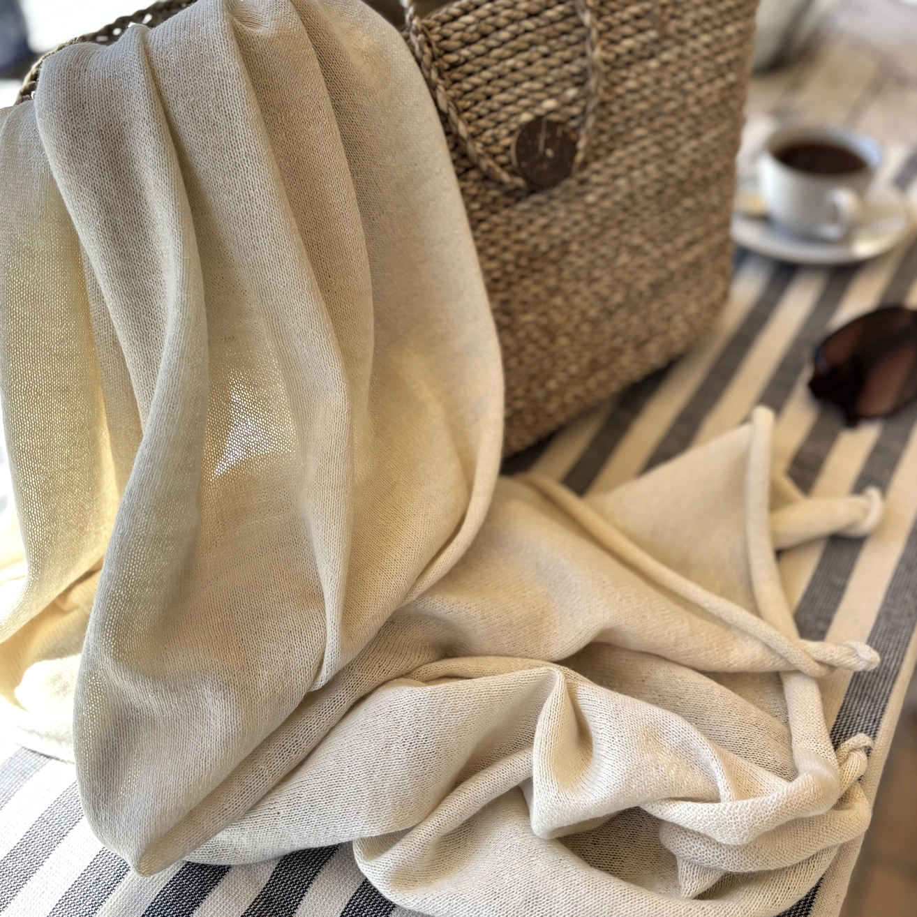 A draped cream-colored linen shawl styled on a striped tablecloth next to a woven straw bag, sunglasses, and a coffee cup.