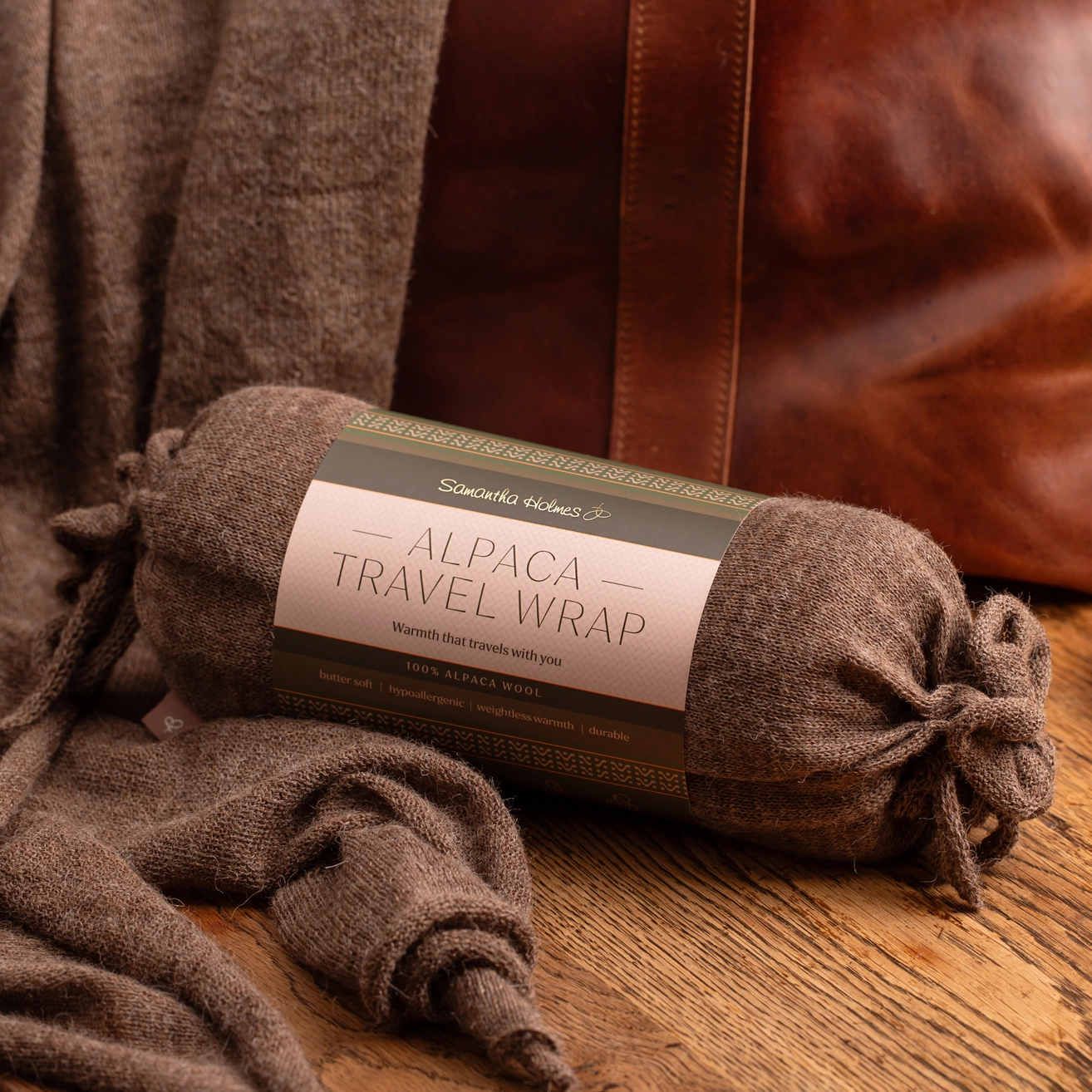 A rolled brown alpaca travel wrap with a paper label styled on a wooden surface with a brown leather bag in the background.