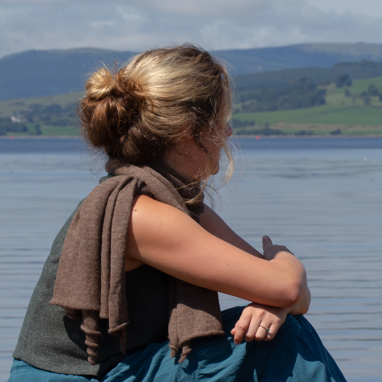 A woman in a gray top and teal skirt sits by a lake with a brown knit wrap draped over her shoulder, with hills in the background.
