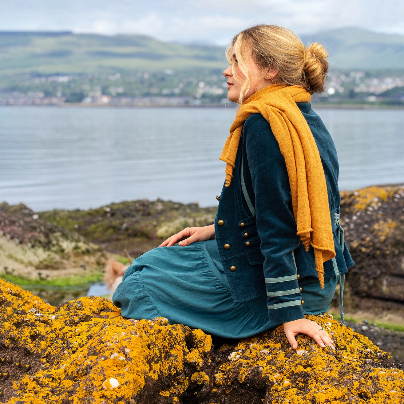 A woman sits on a mossy rock by the water, wearing a teal dress with gold buttons and a mustard yellow scarf, with a coastal town and green hills in the background.