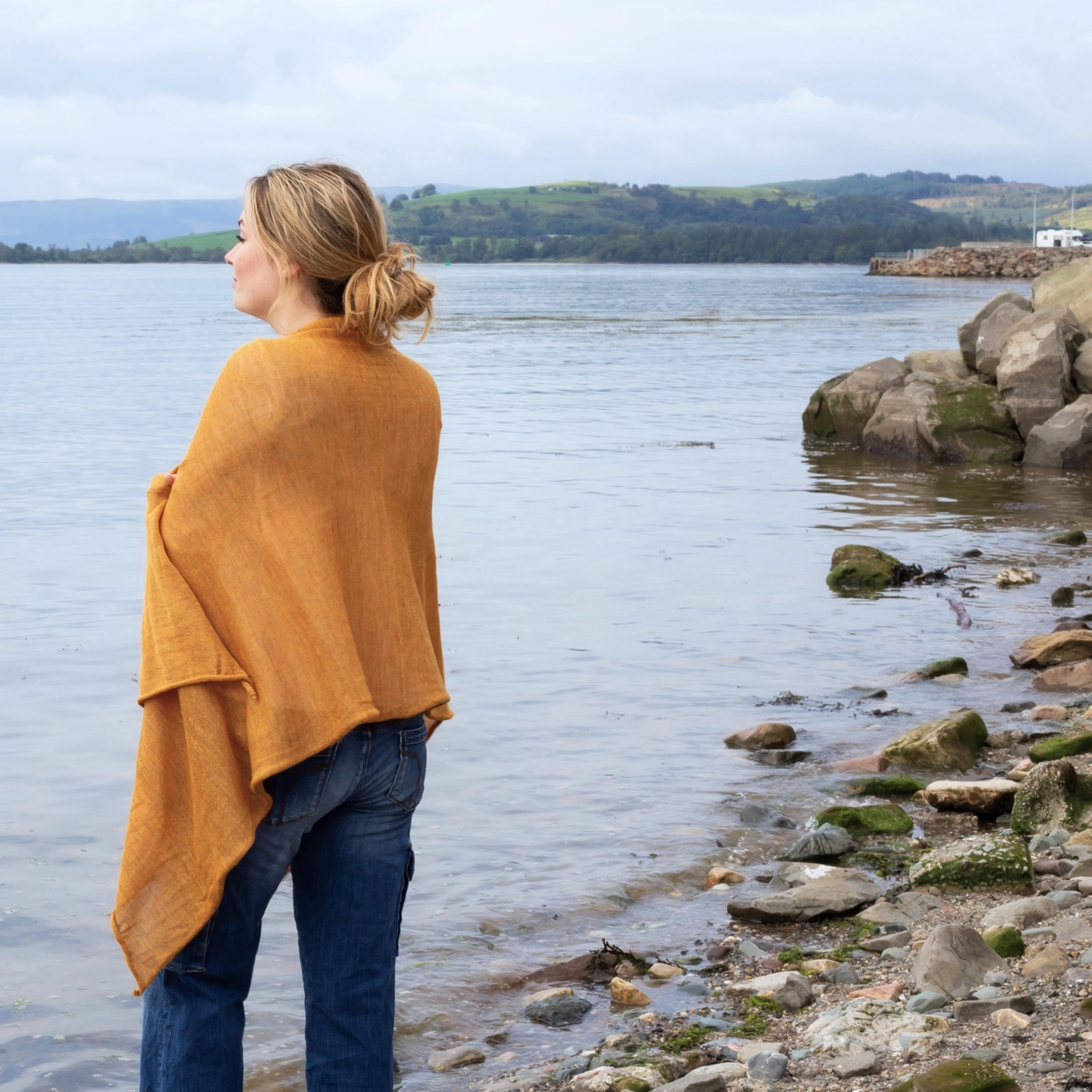 A woman stands on a rocky shoreline by the water, wearing a mustard yellow wrap over blue jeans, with a calm lake and distant green hills under a cloudy sky.