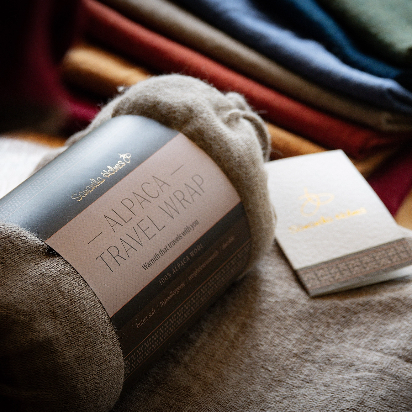 A close-up of a folded beige alpaca travel wrap with a paper label placed on top of other folded colorful textiles.