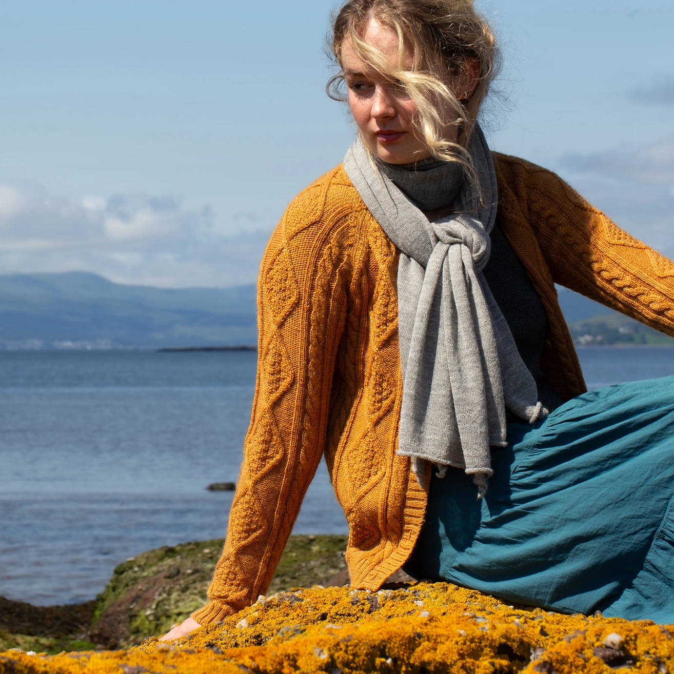 A woman outdoors by the water wearing a chunky cable-knit mustard yellow cardigan, a light gray knit scarf, and a teal skirt, sitting on a rocky shore covered in bright yellow flowers.