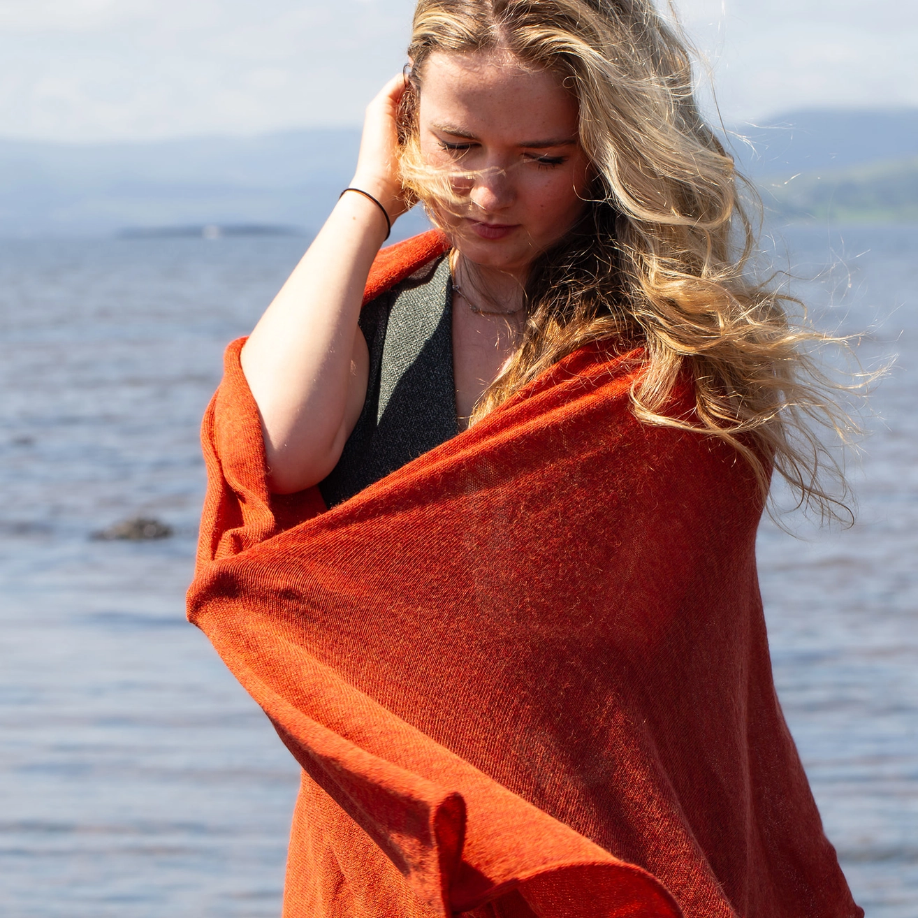 A woman standing outdoors by the water, wrapped in a large rust-orange knit wrap. She has long blonde hair and is wearing a dark gray top underneath.