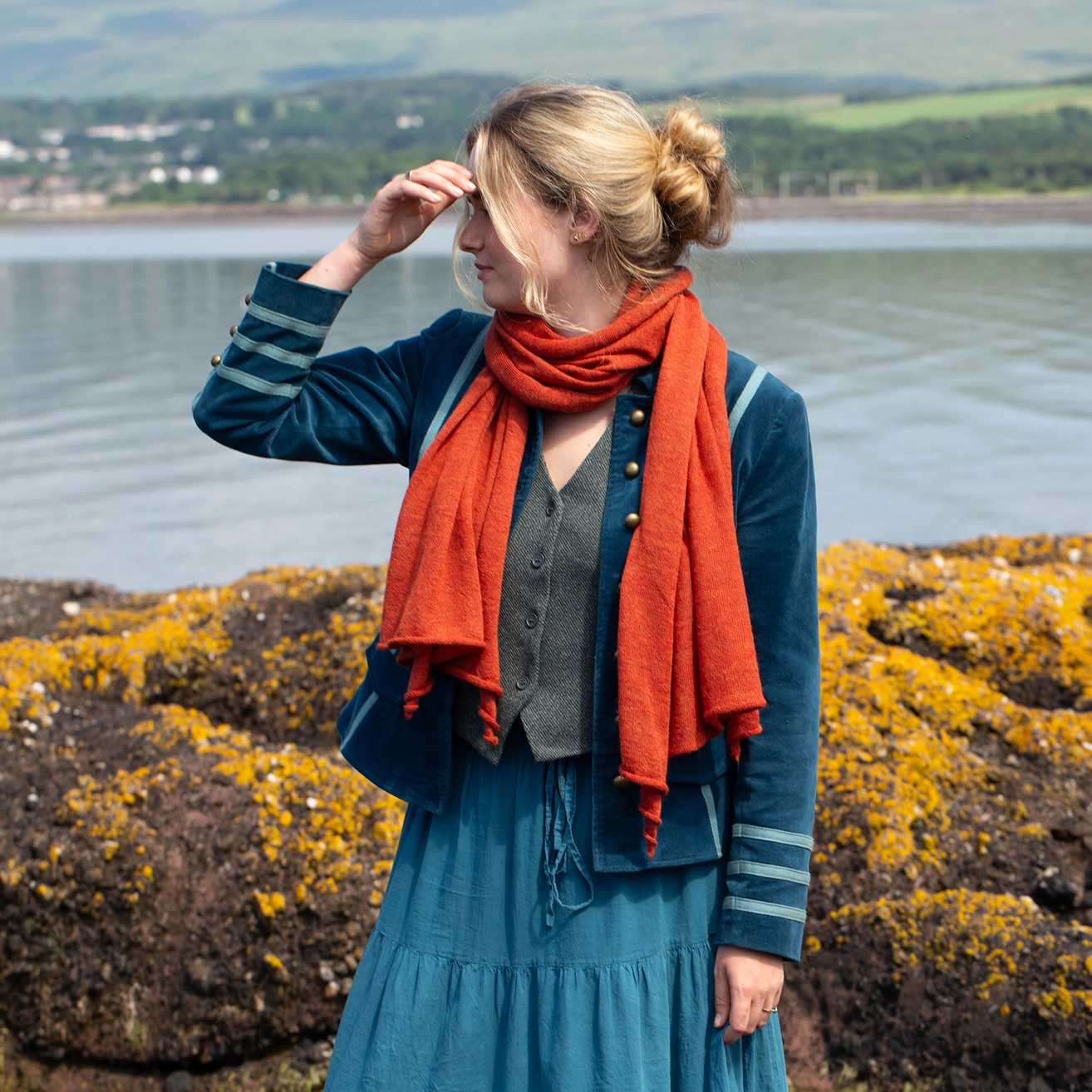A woman standing outdoors by the water wearing a teal dress, blue jacket with striped sleeves, gray vest, and a bright orange-red scarf, with yellow-brown rocks and distant green hills behind her.