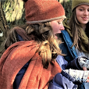 Two people wearing winter clothing and hats, one with an orange scarf and the other with a green hat and scarf, standing close to each other outdoors.