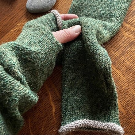 Green knitted gloves being worn on a wooden surface with a stone and in the background.