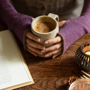 Person holding a mug of coffee on a wooden table with a book and candle in the background