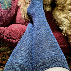Blue striped socks worn on a person's feet, sitting on a fluffy beige cushion with a red pillow.