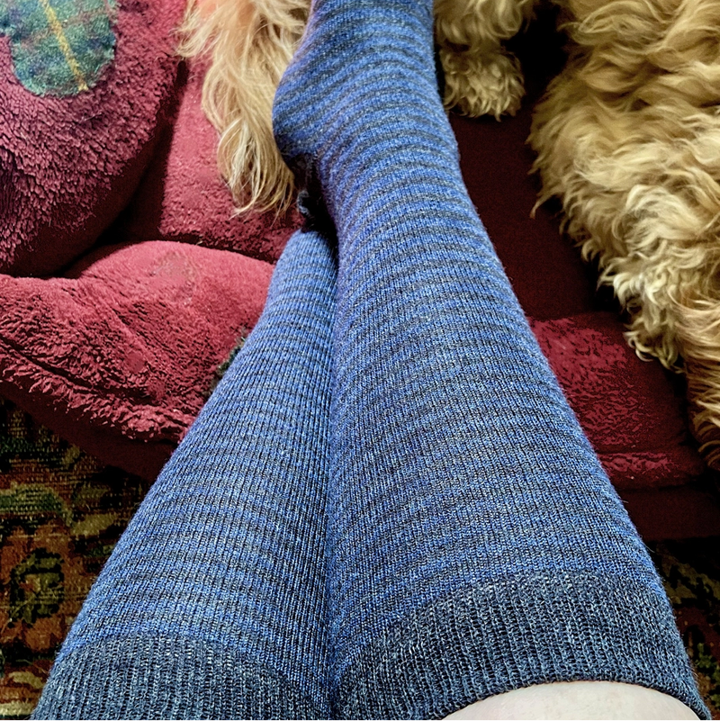 Blue striped socks worn on a person's feet, sitting on a fluffy beige cushion with a red pillow.